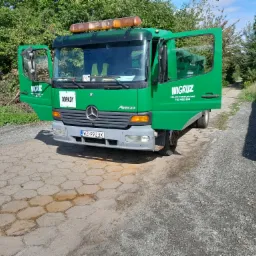 Zielona ciężarówka Mercedes Atego z otwartymi drzwiami, z napisem 'WIGRUZ' i numerem telefonu, zaparkowana na brukowanej powierzchni w słoneczny dzień. Na przedniej szybie widoczna naklejka 'ODPADY'.