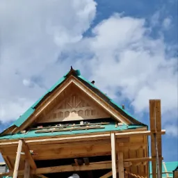 Konstrukcja drewniana domu w trakcie budowy, widoczna więźba dachowa z elementami dekoracyjnymi, zabezpieczona folią, na tle błękitnego nieba z chmurami.