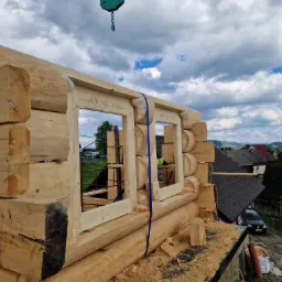 Wznoszenie drewnianego domu z bali: surowe ściany z wyciętymi otworami okiennymi z dekoracyjnymi ramami, dźwig budowlany w tle, widok na wiejską zabudowę i zachmurzone niebo.