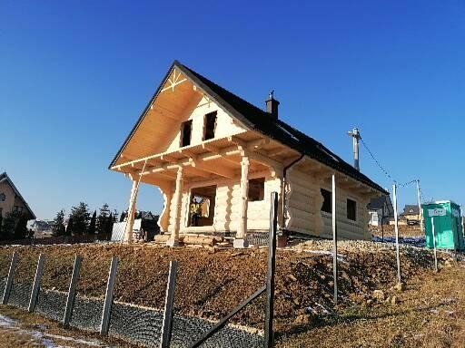 Surowy drewniany dom w trakcie budowy, widoczne bale, niezadaszony taras, czarne pokrycie dachowe, teren wokół domu w trakcie przygotowywania, błękitne niebo.