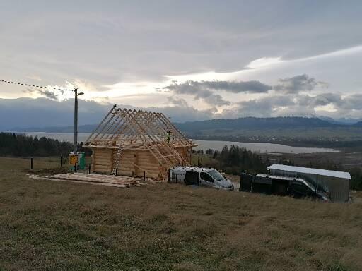 Budowa domu z drewnianych bali, widoczna konstrukcja dachu, na tle górskiego krajobrazu z jeziorem, samochód dostawczy i barak na działce.