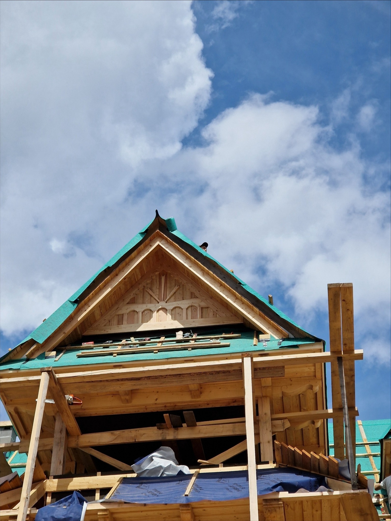 Konstrukcja drewniana domu w trakcie budowy, widoczna więźba dachowa z elementami dekoracyjnymi, zabezpieczona folią, na tle błękitnego nieba z chmurami.