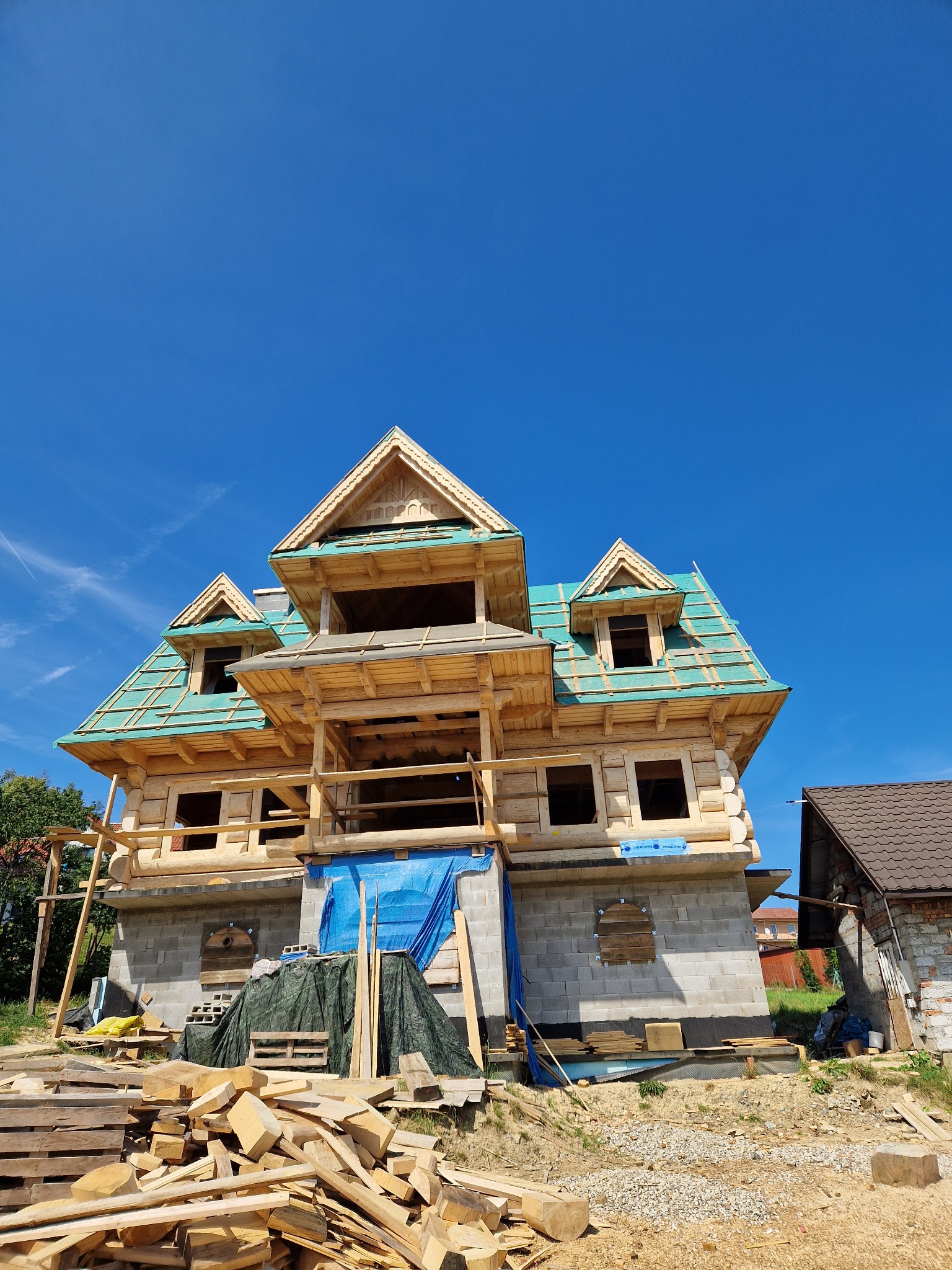 Budowa domu z bali w stanie surowym otwartym, widoczne elementy konstrukcyjne dachu, niebieska plandeka zabezpieczająca, stos drewna na pierwszym planie, błękitne niebo w tle.
