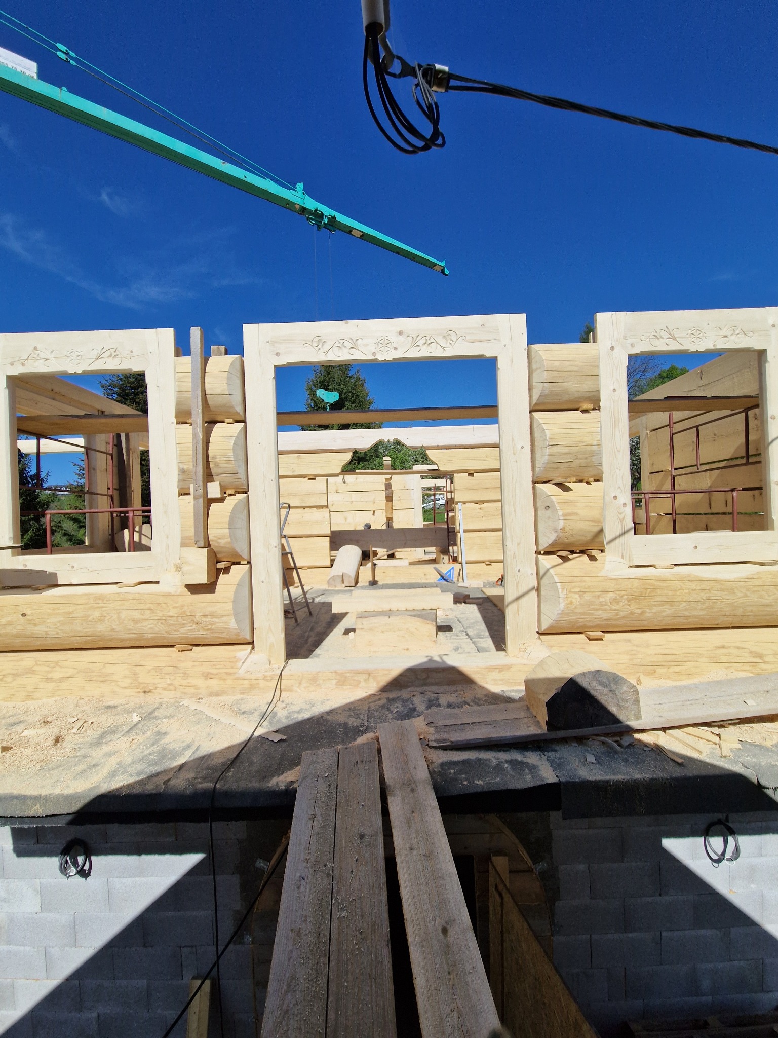 Surowa konstrukcja domu z bali z ozdobnymi wycięciami w drewnie okiennic, widoczny dźwig budowlany i fundamenty z bloczków betonowych.