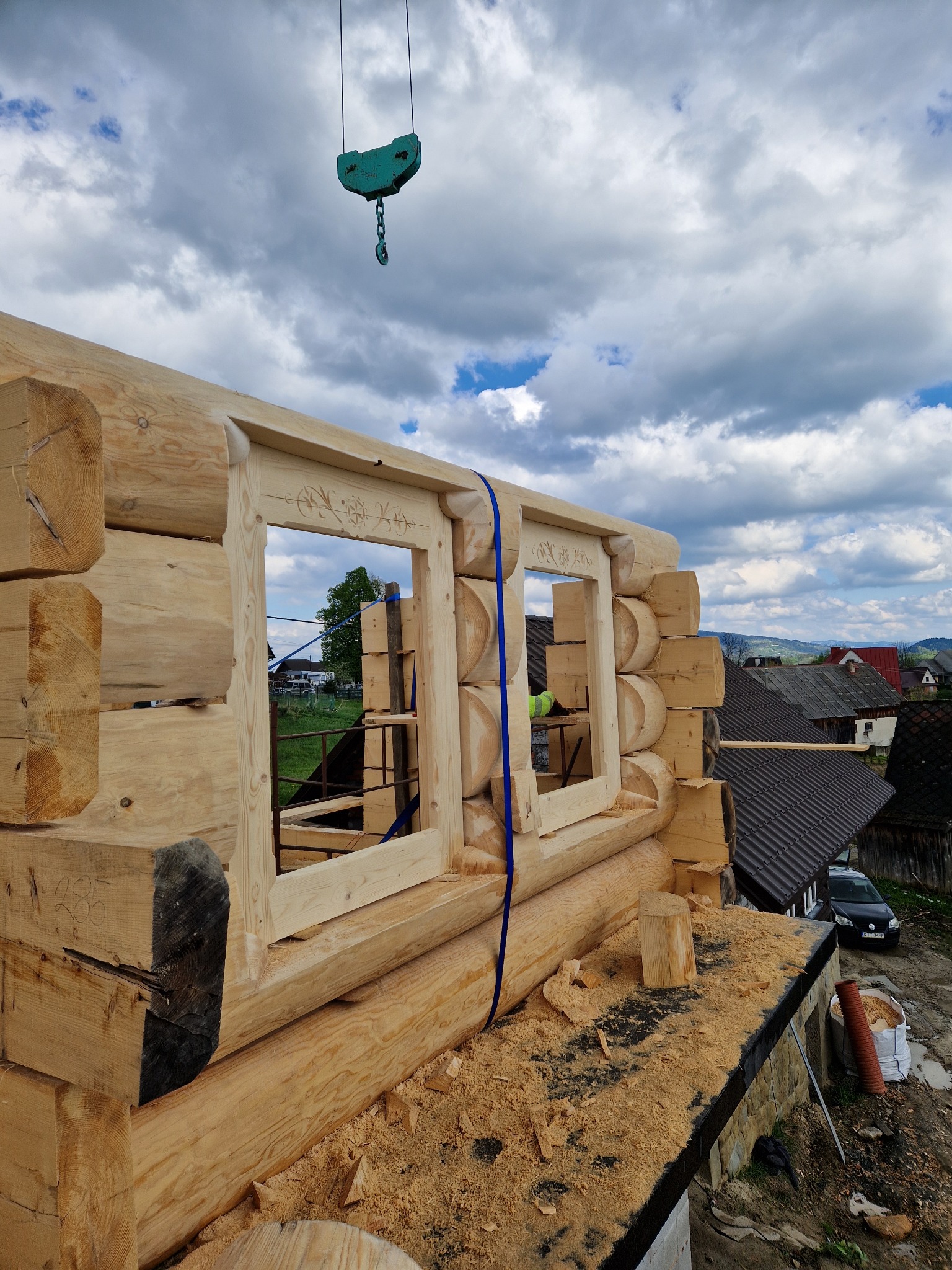 Wznoszenie drewnianego domu z bali: surowe ściany z wyciętymi otworami okiennymi z dekoracyjnymi ramami, dźwig budowlany w tle, widok na wiejską zabudowę i zachmurzone niebo.