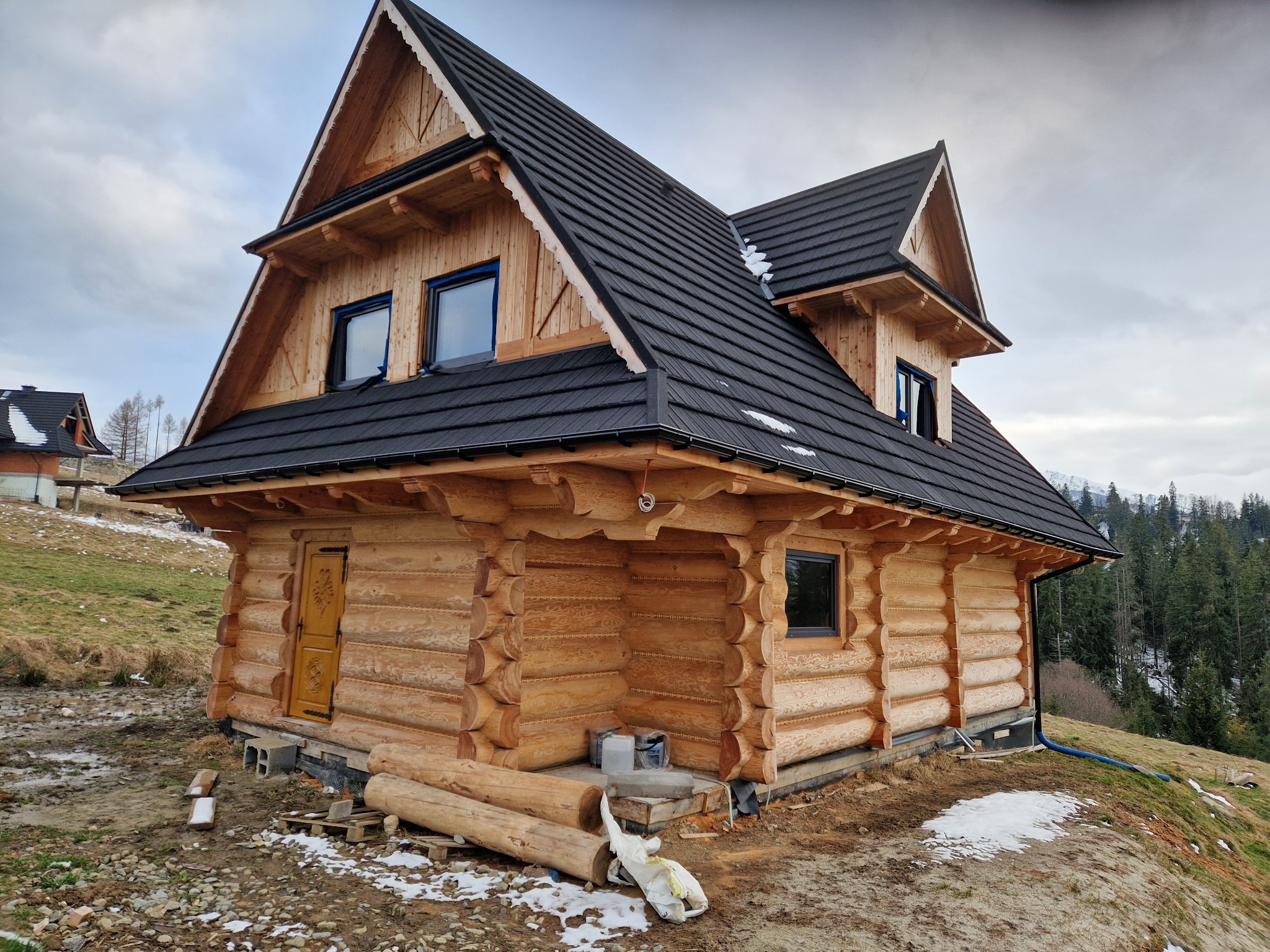 Dwukondygnacyjny dom z bali w stylu góralskim z ciemnym dachem, widok z boku na tle zalesionego wzgórza, w trakcie budowy, z leżącymi obok kłodami drewna.