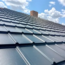 Nowoczesne, grafitowe pokrycie dachowe z blachy trapezowej, widok z góry, na tle błękitnego nieba z białymi obłokami, z kominem z czerwonej cegły w tle.