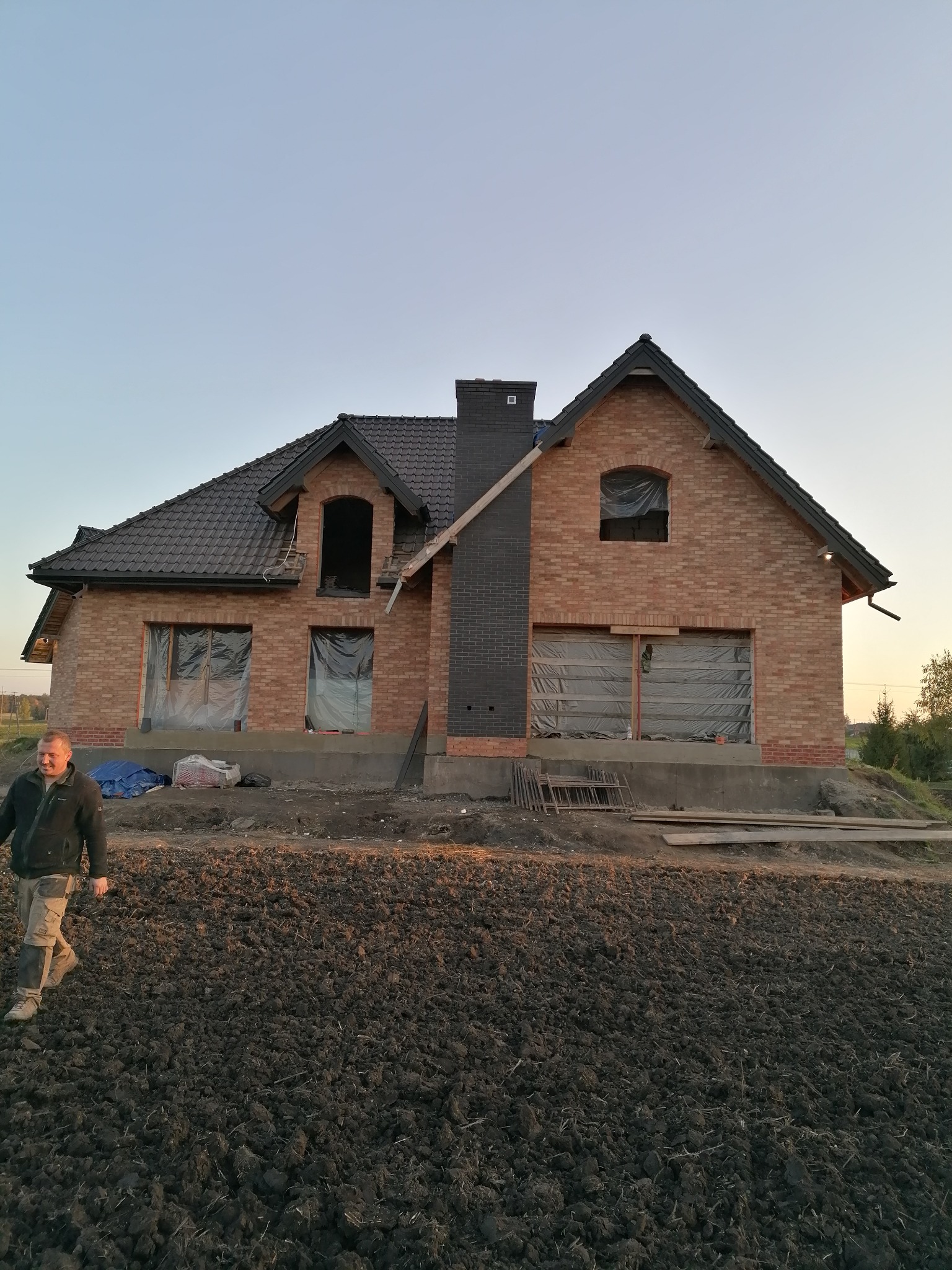 Niedokończony dom z cegły z czarnym dachem i kominem, okna zabezpieczone folią, pracownik w ubraniu roboczym przechadza się po polu w tle.