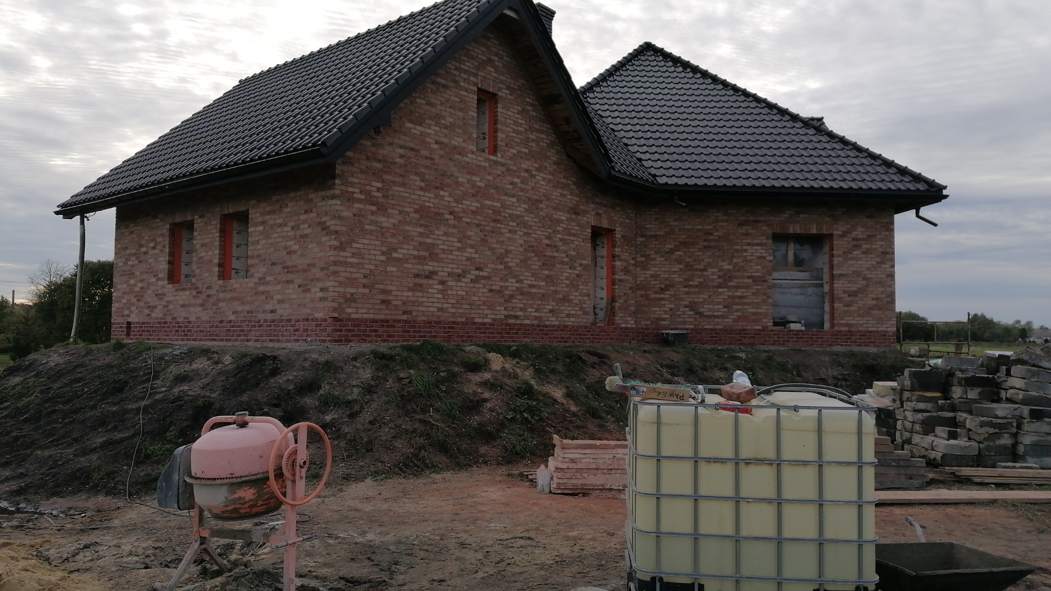 Wznoszony dom z czerwonej cegły z ciemnym dachem, widoczne otwory okienne i drzwiowe, na pierwszym planie betoniarka i zbiornik na wodę, teren budowy.
