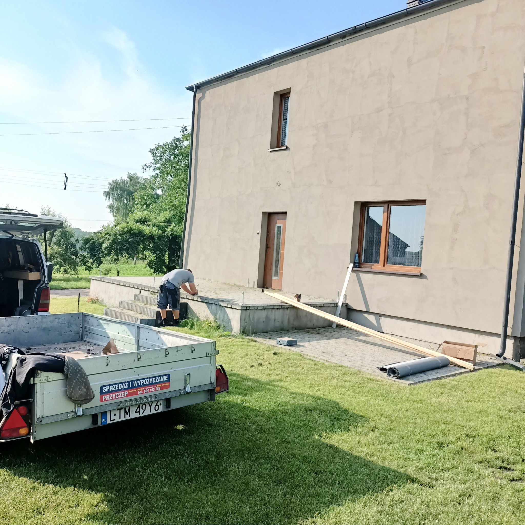 Pracownik przygotowuje się do montażu listew przypodłogowych na elewacji budynku, przyczepa z narzędziami i materiałami w tle.