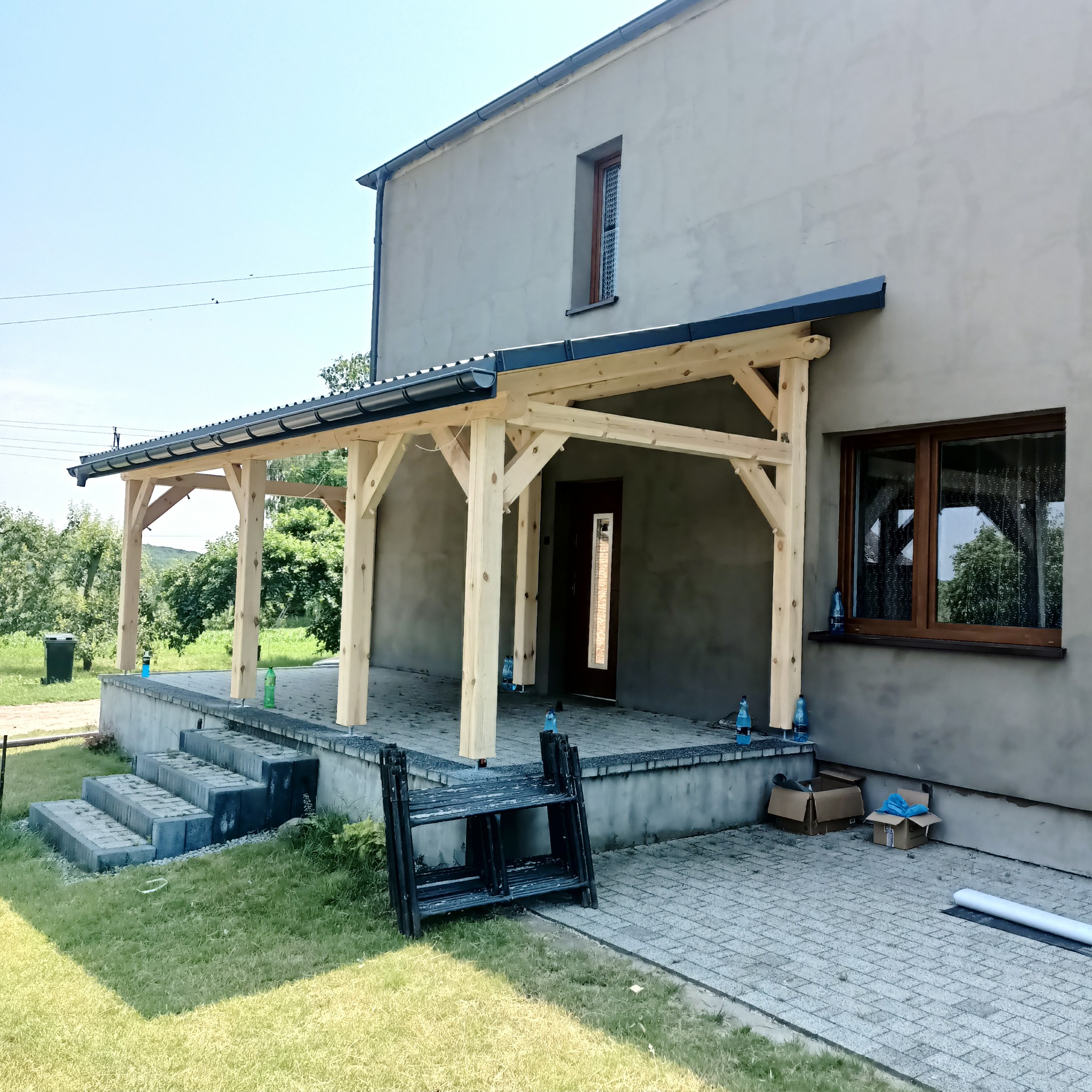 Drewniana konstrukcja z zadaszeniem w trakcie budowy przy szarym domu, widoczne schody wejściowe, trawnik i narzędzia pracy.