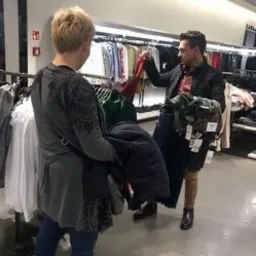 Mężczyzna w Ostródzie pomaga kobiecie wybrać ubrania w sklepie odzieżowym, trzymając kilka sztuk na wieszakach.