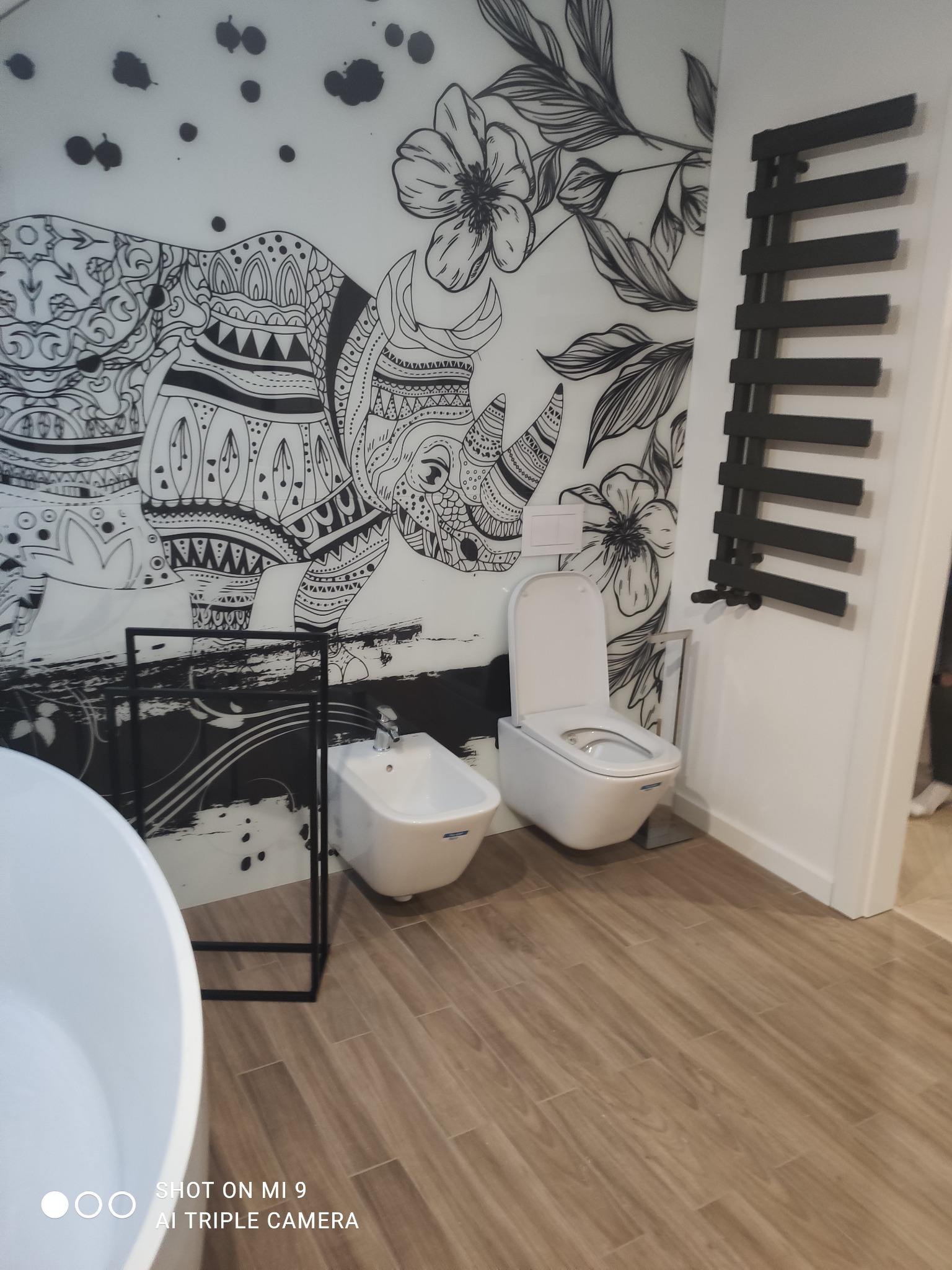 Nowoczesna łazienka po remoncie: biała ceramika (toaleta, bidet), grzejnik drabinkowy na tle ściany z dekoracyjnym motywem słonia i kwiatów. Drewnopodobne płytki podłogowe.