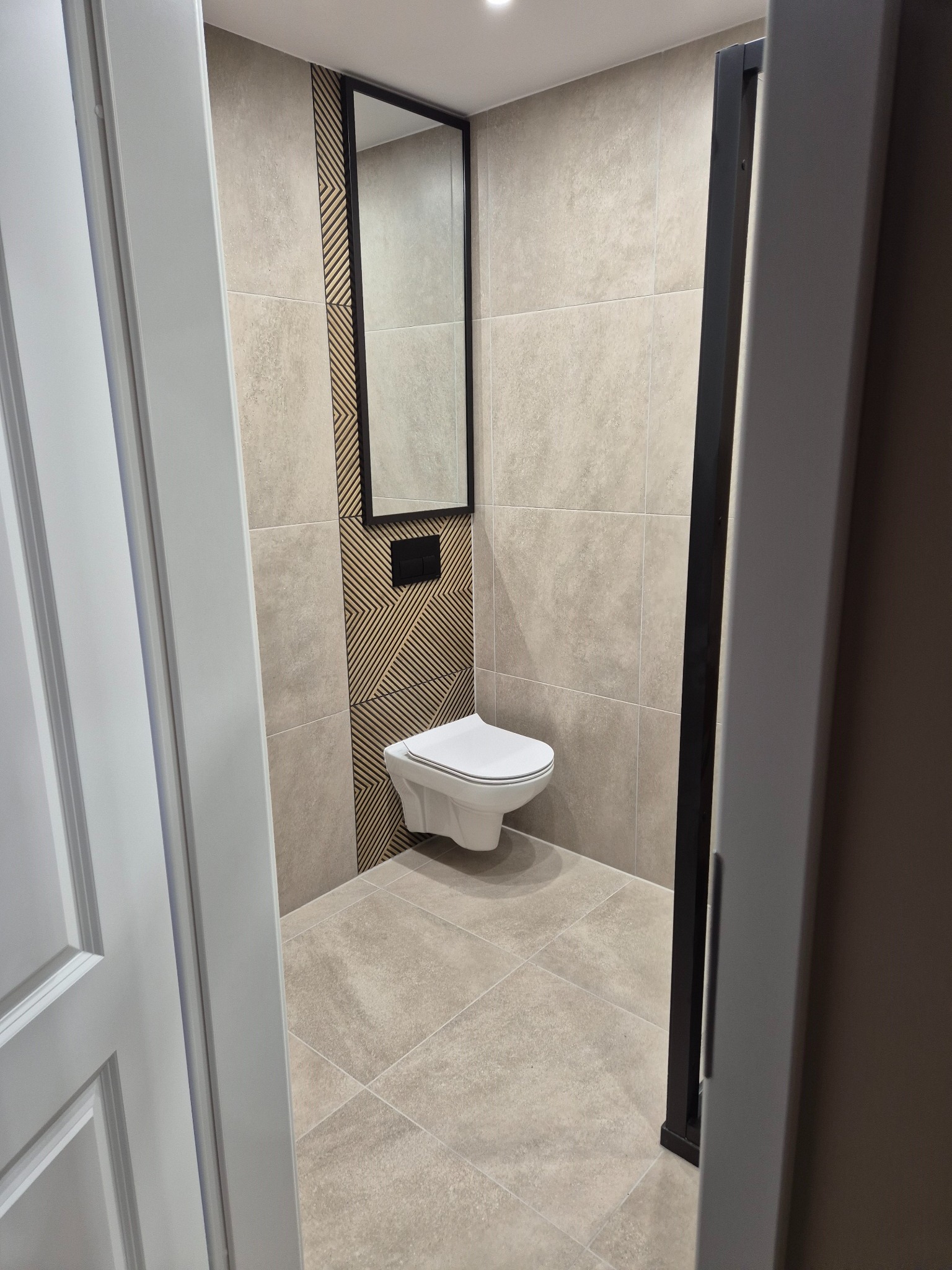 Wykończona łazienka z beżowymi płytkami, wiszącą toaletą i lustrem w czarnej ramie, widoczna zza uchylonych białych drzwi. Dekoracyjna ściana z drewnianymi elementami.