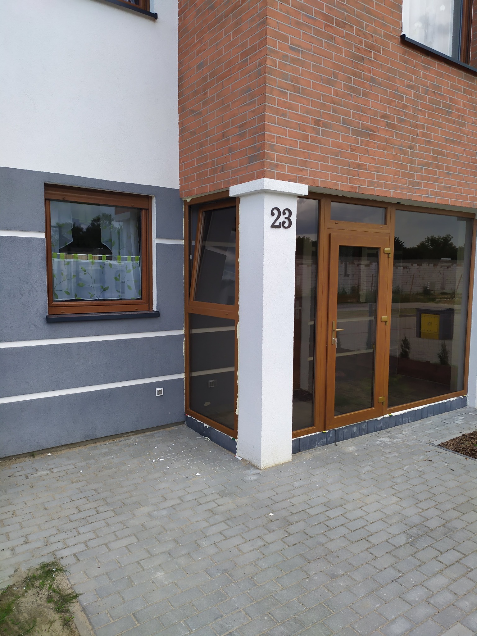 Nowe okna i przeszklone wejście do budynku z numerem 23, widoczne ślady prac wykończeniowych na kostce brukowej.