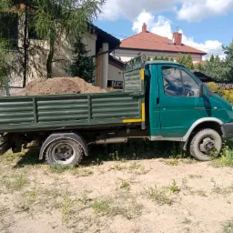 Zielony samochód dostawczy z otwartą skrzynią załadowaną ziemią, zaparkowany na piaszczystym terenie z rzadką roślinnością, w tle domy jednorodzinne pod błękitnym niebem z chmurami.