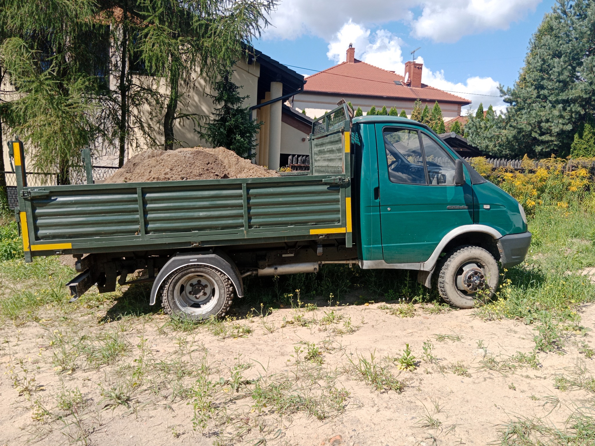 Zielony samochód dostawczy z otwartą skrzynią załadowaną ziemią, zaparkowany na piaszczystym terenie z rzadką roślinnością, w tle domy jednorodzinne pod błękitnym niebem z chmurami.