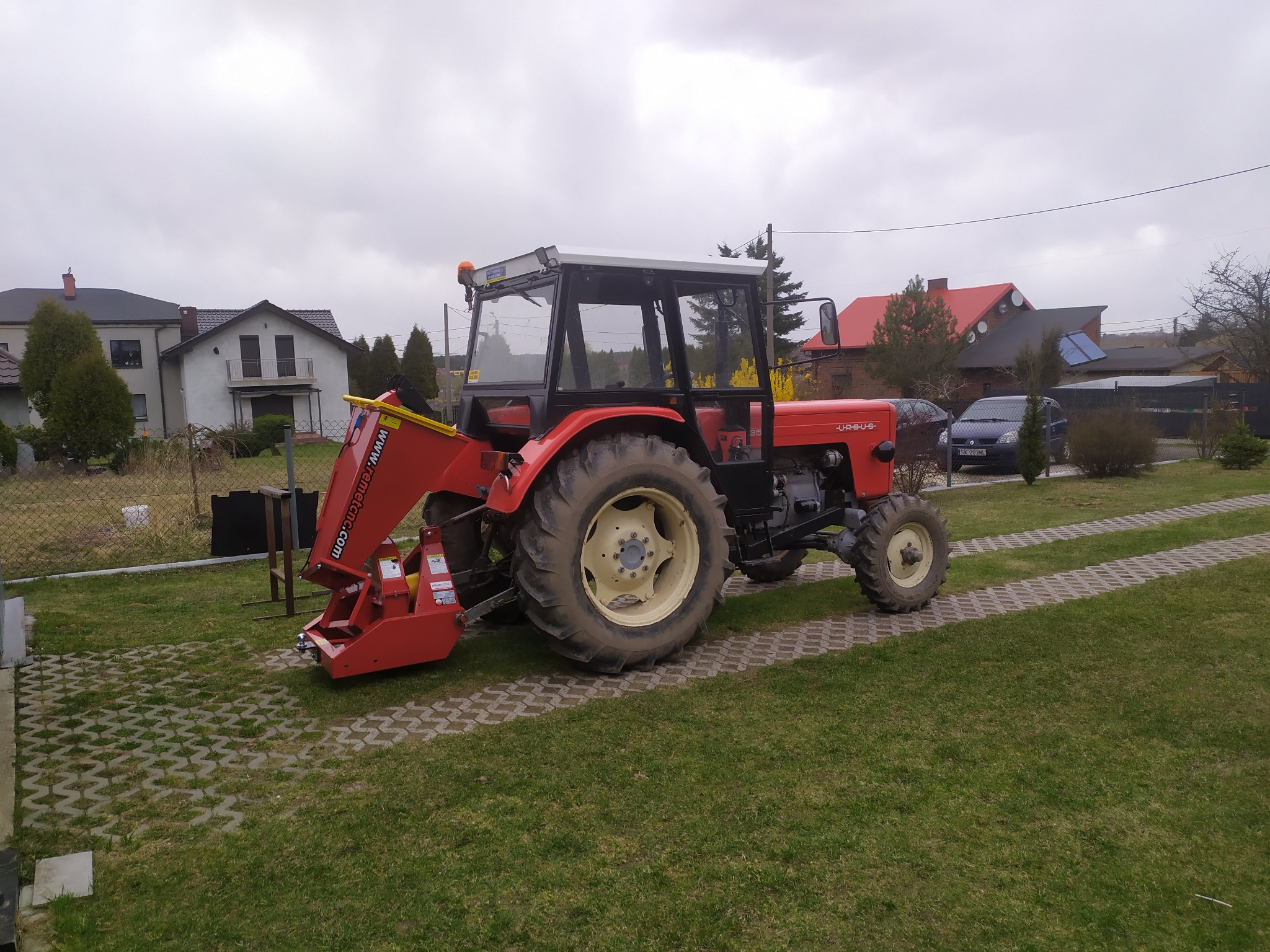 Czerwony traktor Ursus z zamontowanym osprzętem do prac ziemnych stoi na trawniku przed domem z kostki ażurowej.