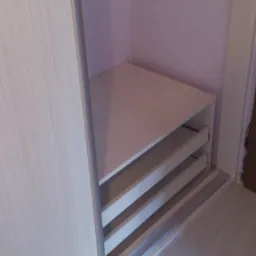 Wnętrze szafy wnękowej z trzema szufladami z jasnego drewna, widoczne prowadnice szuflad i przesuwne drzwi szafy z jasnego drewna na tle fioletowej ściany.