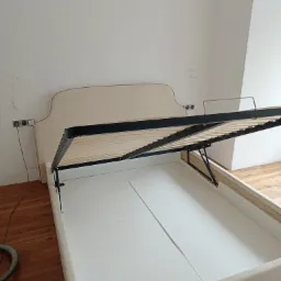 Beżowe łóżko z podnoszonym stelażem i pojemnikiem na pościel, widoczne mechanizmy podnoszące, surowe wykończenie wnętrza, gniazdka elektryczne na ścianie.