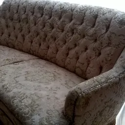 Odrestaurowana sofa w stylu retro z tapicerką w kwiatowy wzór, wykończona ozdobnymi gwoździami, na tle okna z zasłonami.