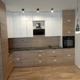 Montaż kuchni: Dolne szafki w odcieniach drewna, górne białe, czarny blat i piekarnik, widoczne gniazdka elektryczne, lampy sufitowe i fragment podłogi w trakcie prac montażowych.