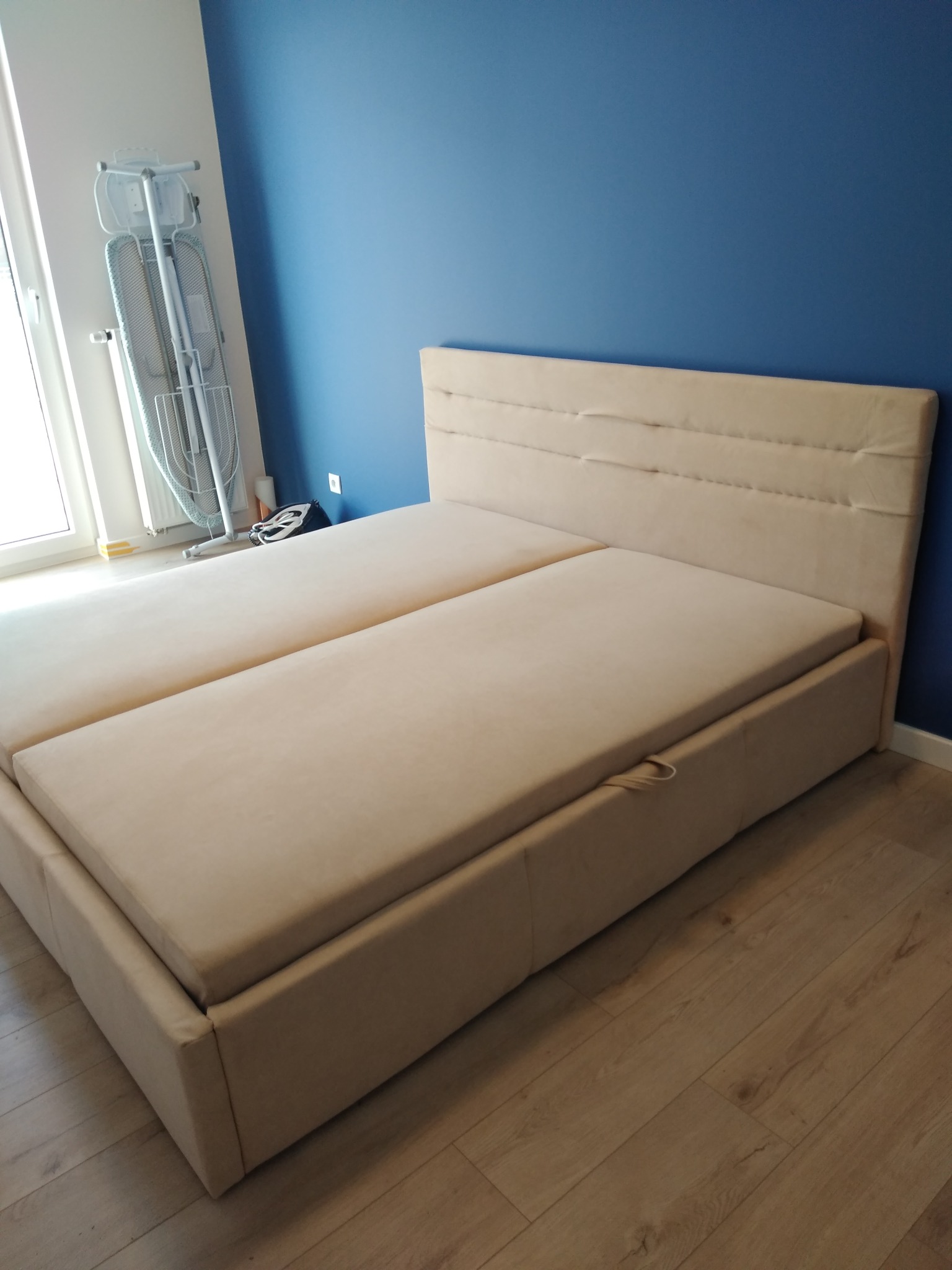 Beżowe, tapicerowane łóżko kontynentalne z pojemnikiem na pościel, widoczne na tle niebieskiej ściany i parkietu, obok okna z deską do prasowania.