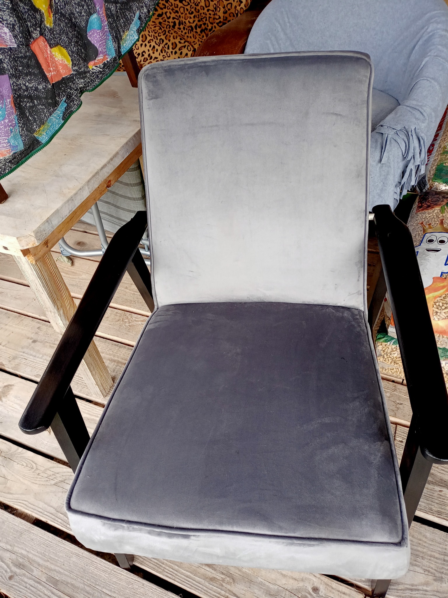 Szare, tapicerowane krzesło z ciemnymi, drewnianymi podłokietnikami, ustawione na drewnianym tarasie, z widocznymi elementami otoczenia w tle.