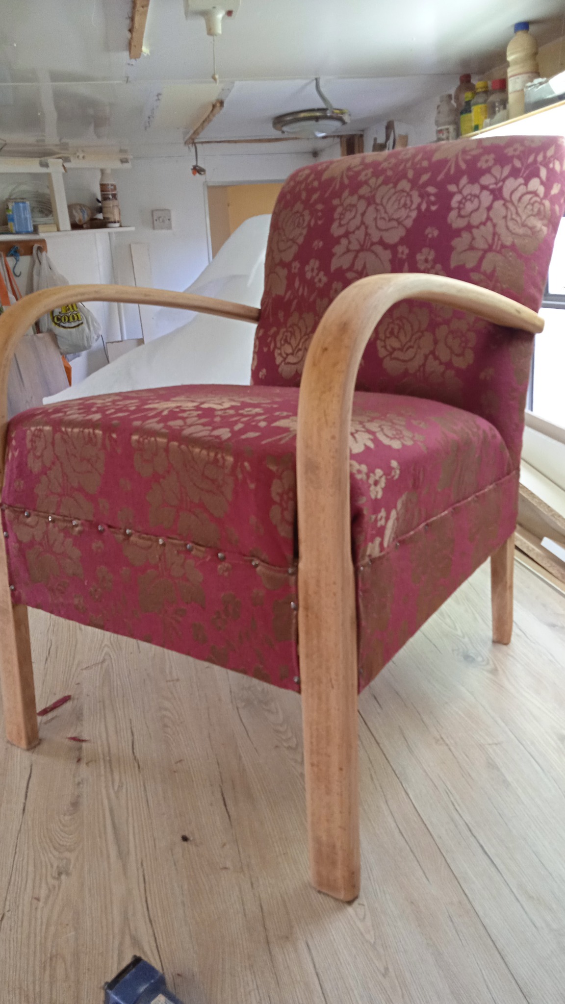 Odrestaurowany fotel z drewnianymi podłokietnikami i bordową tapicerką w złote kwiaty, wykończony ozdobnymi gwoździami, stoi na panelach podłogowych w warsztacie.