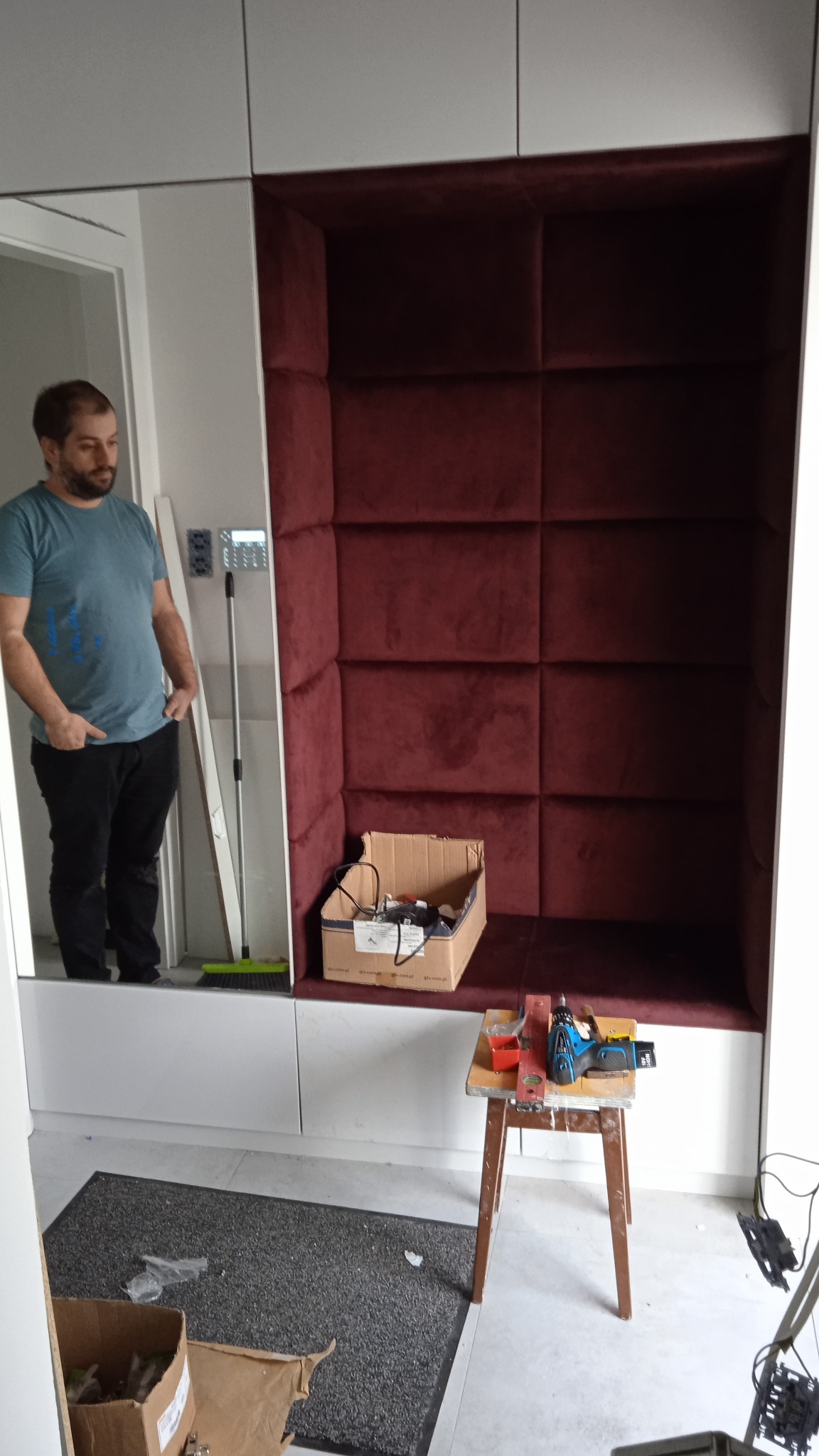 Wnęka z bordowym tapicerowanym obiciem w zabudowie meblowej, widoczny mężczyzna w tle, narzędzia na taborecie i karton z akcesoriami.