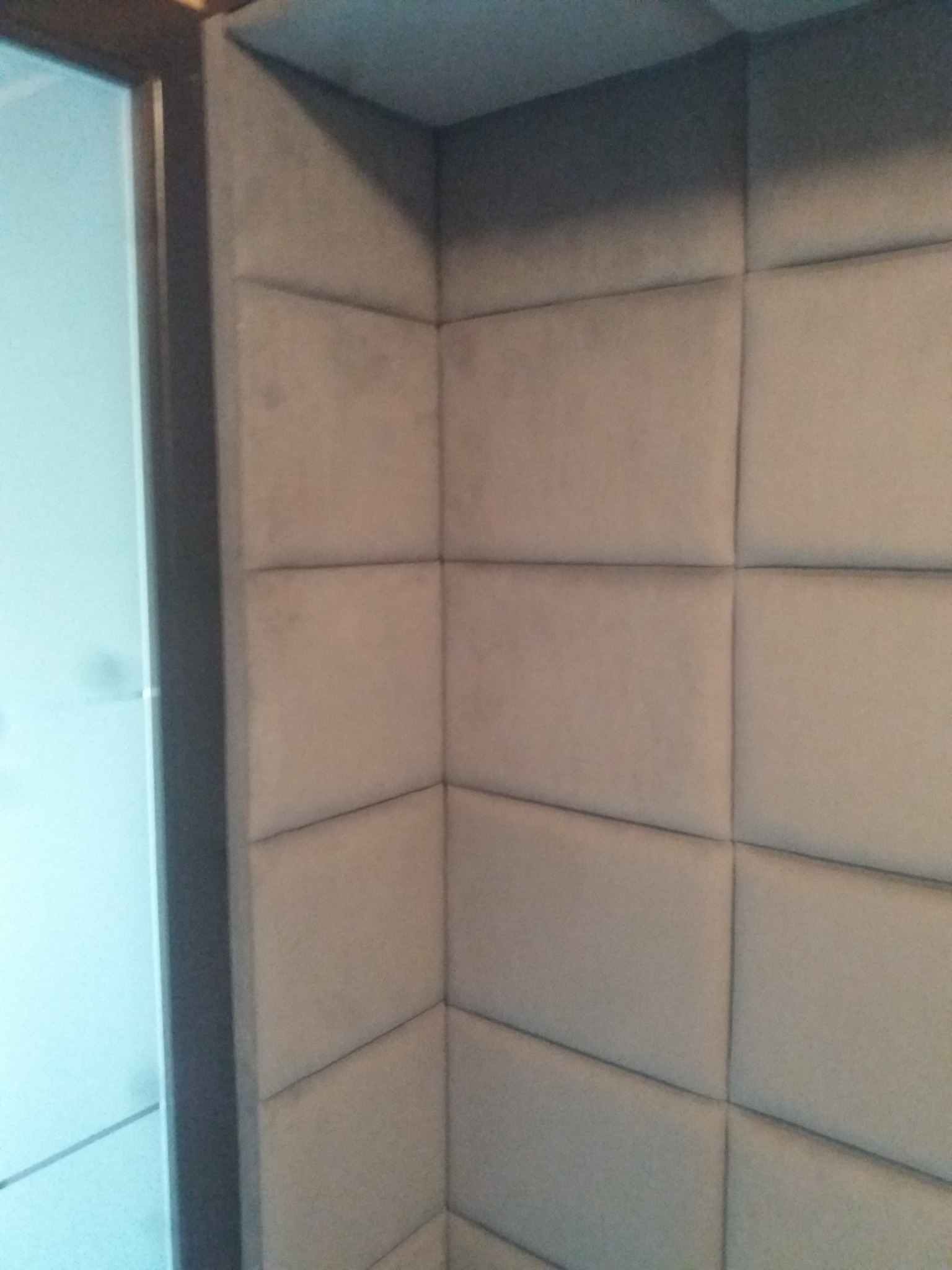 Wykończenie narożnika pokoju panelami tapicerowanymi w jasnym odcieniu szarości, z widocznym fragmentem ościeżnicy drzwiowej po lewej stronie.