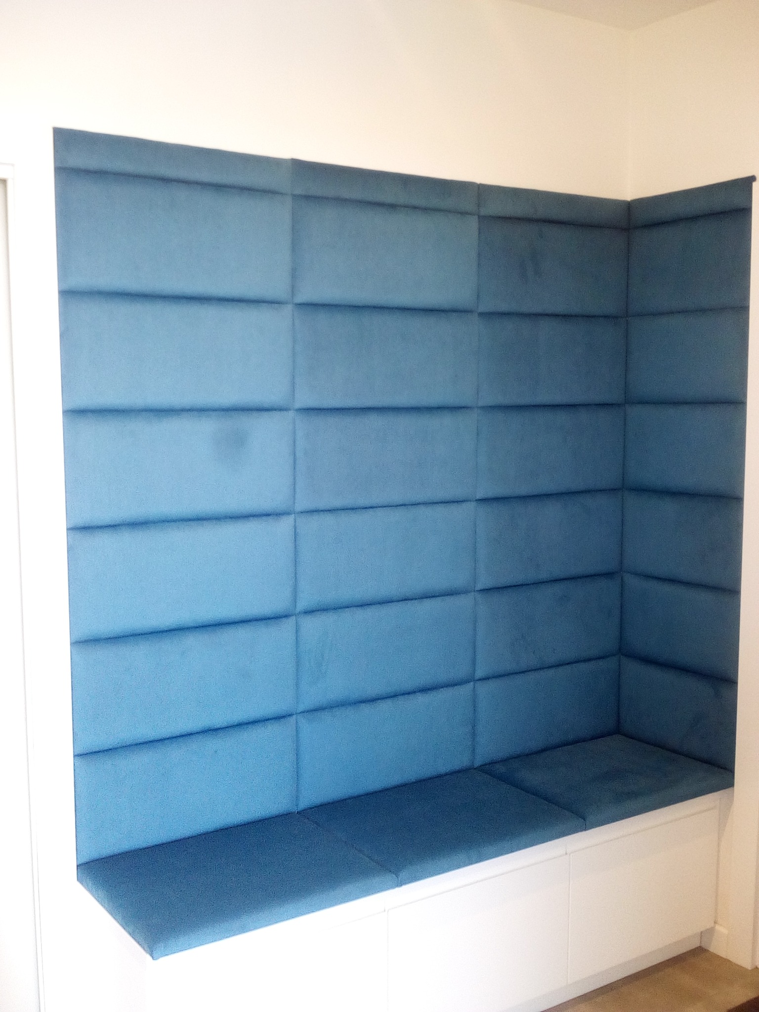 Narożna wnęka z siedziskiem i wysokim, błękitnym panelem tapicerowanym w kwadraty, umieszczona w białej ramie.