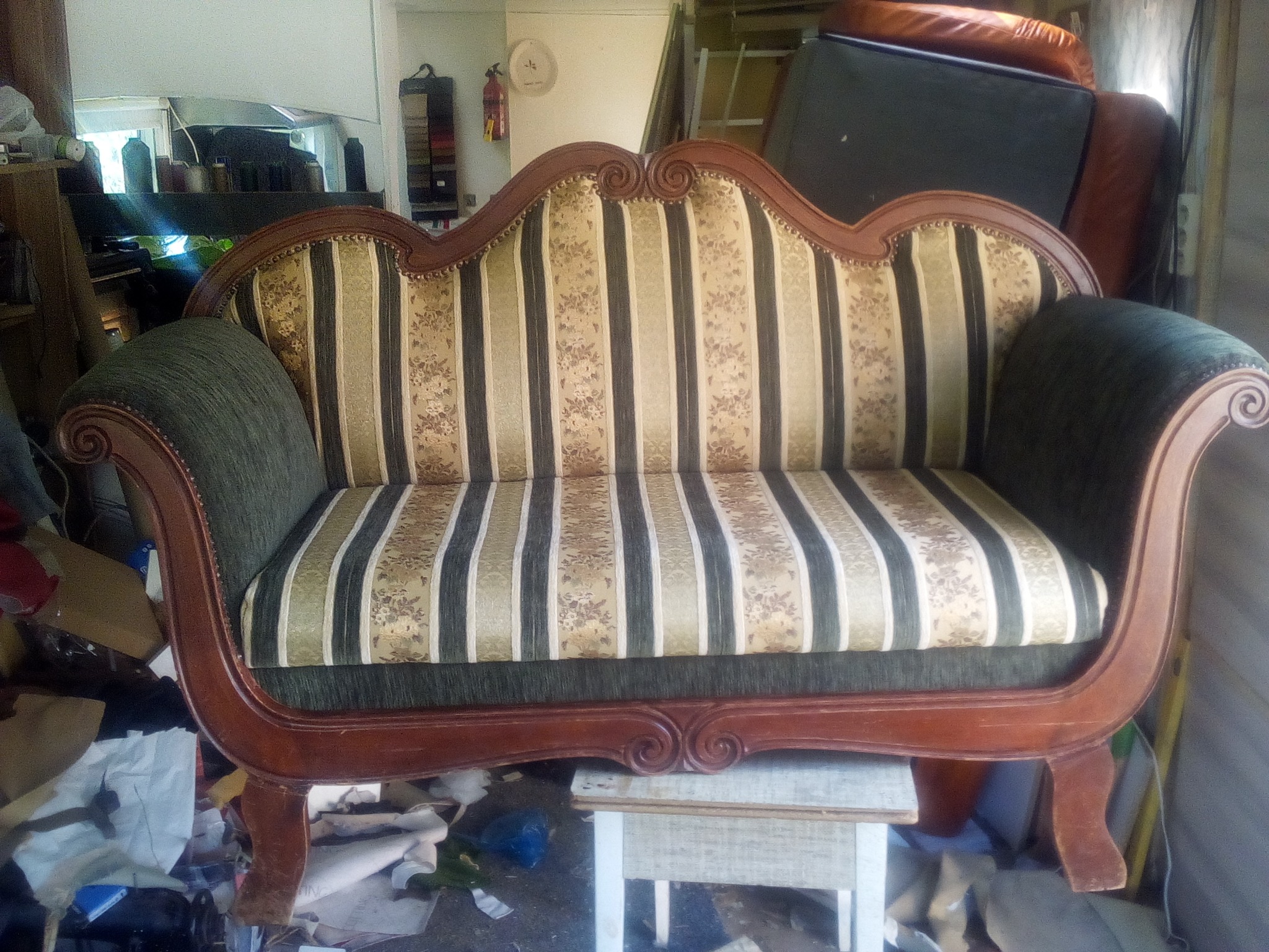 Odrestaurowana sofa w stylu retro z drewnianą ramą, tapicerowana w pasy z motywem kwiatowym, umieszczona na małym stoliku w warsztacie.