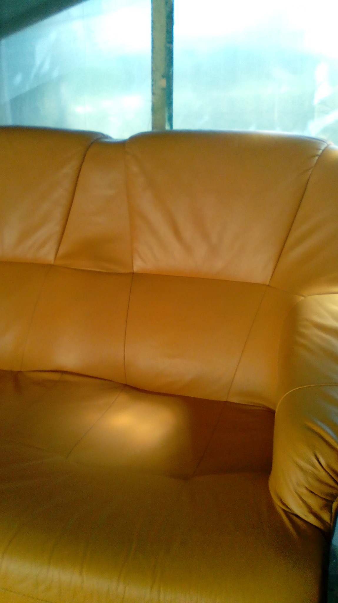 Fragment sofy z jasnej skóry, widoczne przeszycia i zaokrąglone ramię, oświetlone naturalnym światłem wpadającym przez okno z niebieską szybą.