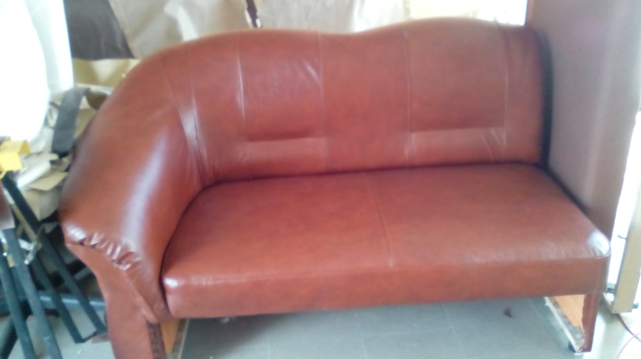 Brązowa, dwuosobowa sofa z ekoskóry, z charakterystycznym zaokrąglonym podłokietnikiem po lewej stronie, sfotografowana w warsztacie tapicerskim, widoczne elementy konstrukcyjne i narzędzia w tle.