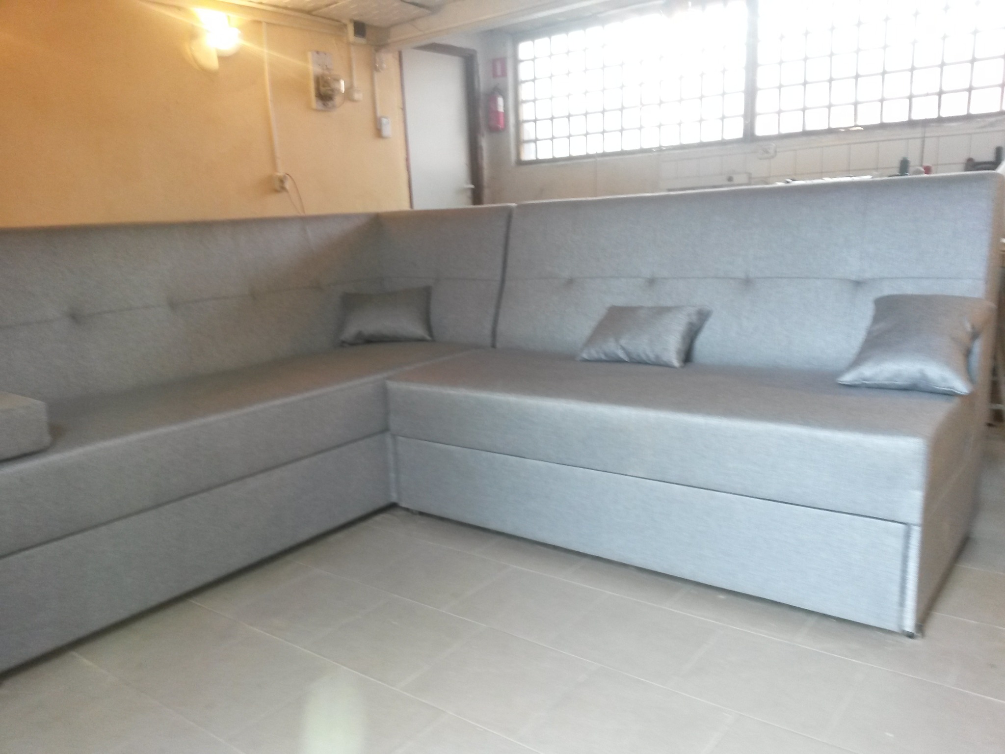 Szary narożnik z poduszkami w jasnym pomieszczeniu z oknem w kratkę, widoczne szwy i guziki na oparciu.