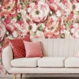 Jasnoszara sofa z dwoma poduszkami w odcieniach różu i bordo na tle ściany pokrytej tapetą w kwiaty w stylu vintage.