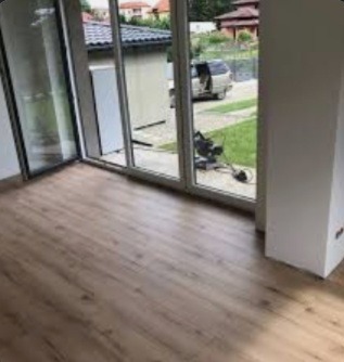 Pusty pokój po remoncie z nową, jasną podłogą z paneli, duże okno przesuwne z widokiem na ogród i samochód dostawczy z otwartymi drzwiami, sugerujący zakończenie prac.