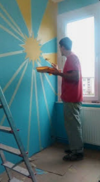 Mężczyzna w czerwonej koszulce maluje ścianę na niebiesko z motywem słońca, używając pędzla i kuwety, drabina oparta o ścianę, widok z boku, pokój w trakcie odświeżania.