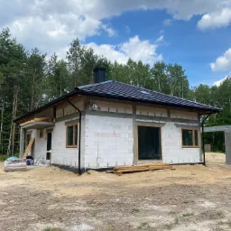 Surowy stan domu jednorodzinnego z białych bloczków, czarny dach z kominem, widoczne drewniane ramy okienne, teren wokół w trakcie przygotowania, las w tle.