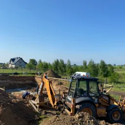 Wykonanie wykopu pod fundamenty domu jednorodzinnego z użyciem koparko-ładowarki Case 580ST, widoczne szalunki i pracownik w wykopie, w tle zabudowa jednorodzinna i błękitne niebo.