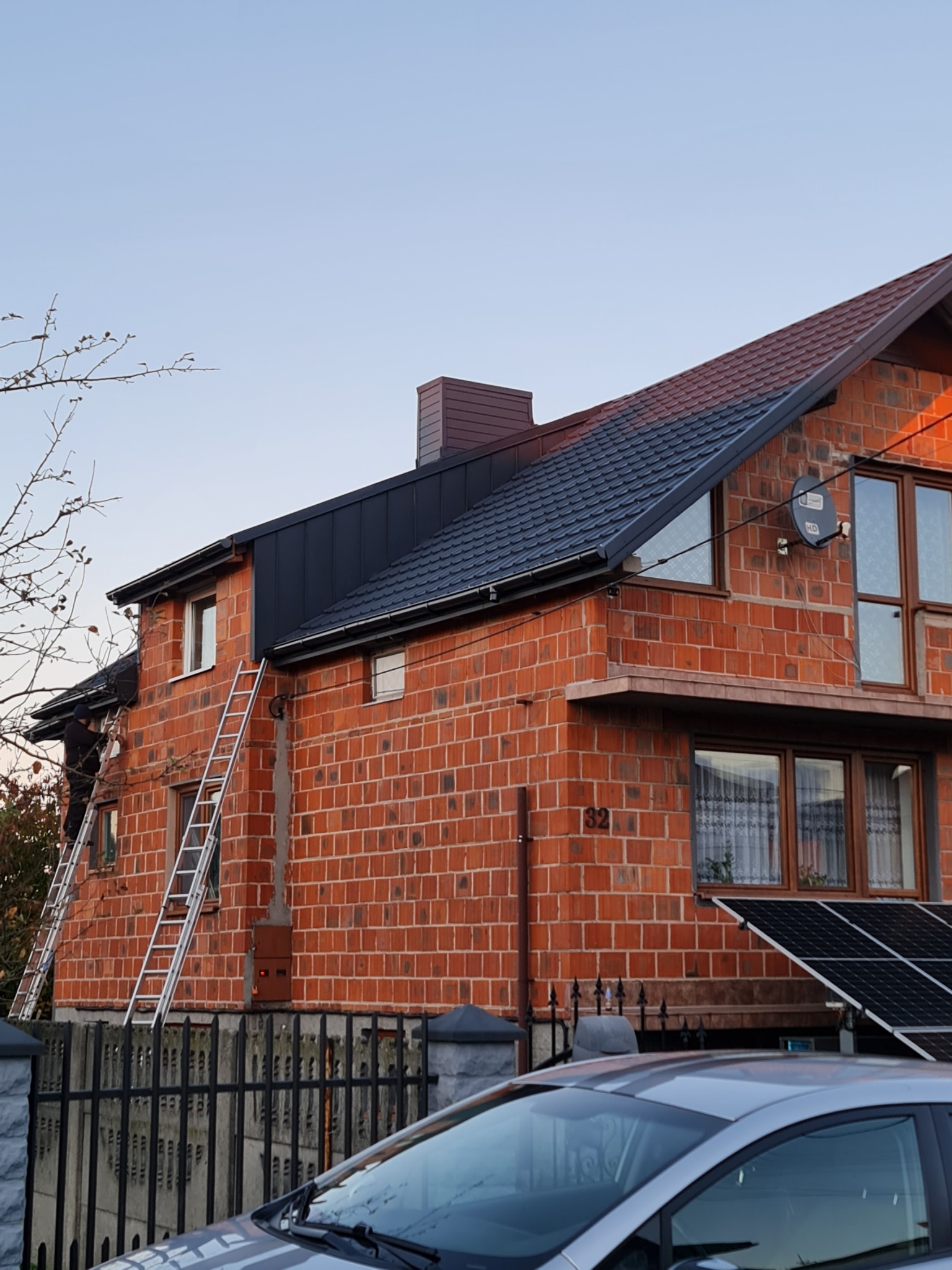 Murarz na drabinie, w trakcie prac na dachu dwupiętrowego domu z czerwonej cegły, widoczne panele słoneczne, dach pokryty czarną blachodachówką, fragment samochodu na pierwszym planie.