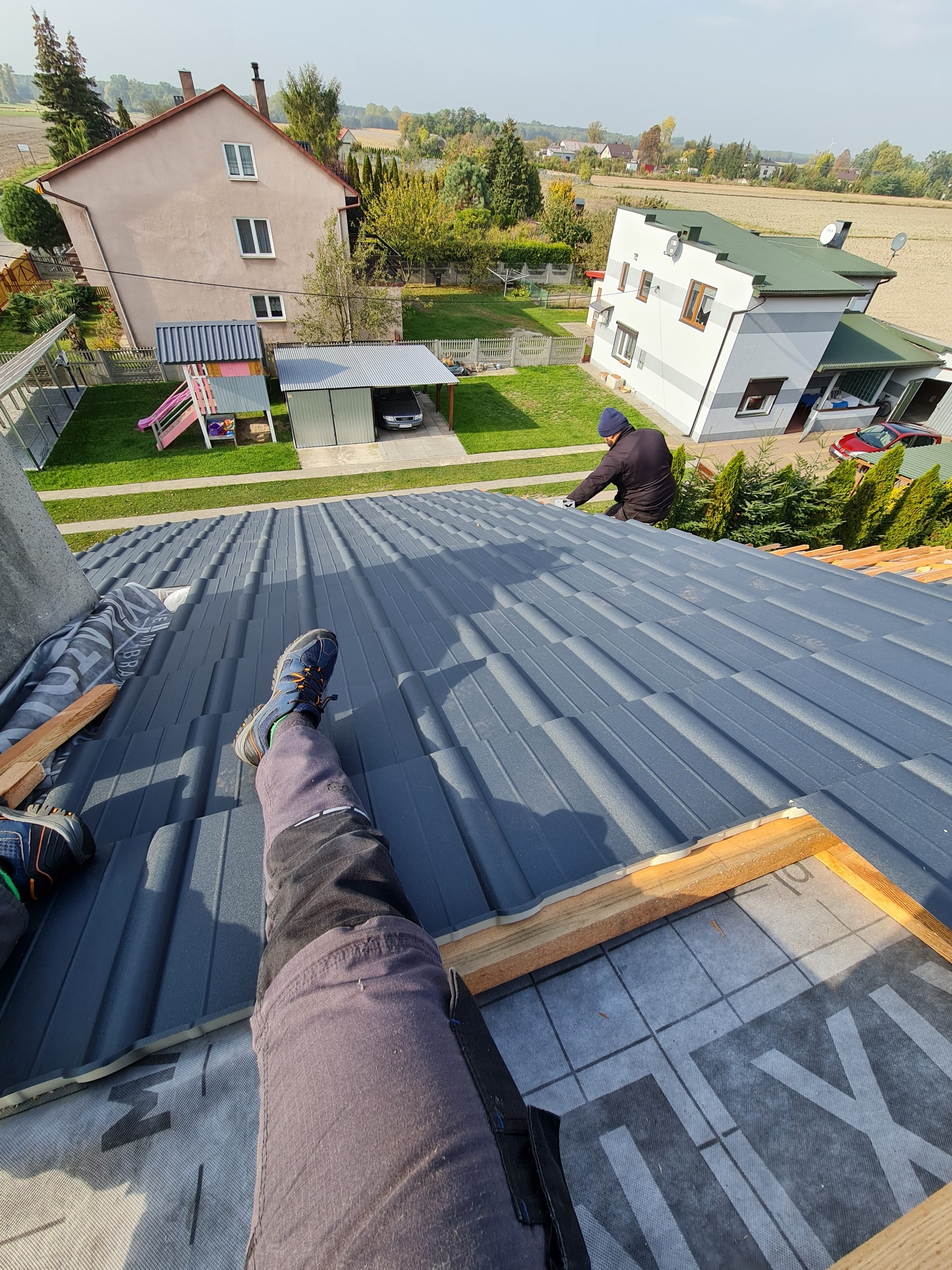 Widok z góry na układanie szarej blachy na dachu domu jednorodzinnego, widoczne fragmenty drewnianej konstrukcji, membrana dachowa i pracownik w oddali oraz nogi fotografa w obuwiu sportowym...