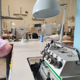 Wnętrze szwalni z kilkoma maszynami do szycia, w tym overlock z czterema nićmi na pierwszym planie, oświetlonym lampą przemysłową, w tle widoczne półki i inne maszyny.