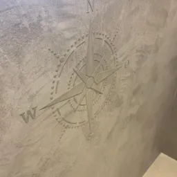 Dekoracyjna ściana z szarym tynkiem strukturalnym i wytłoczonym motywem róży wiatrów z literami N, W, S, O.