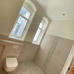 Jasna łazienka z ukośnymi oknami, beżowe płytki na ścianach z dekoracyjnym pasem mozaiki, podwieszana toaleta i mozaika podłogowa.