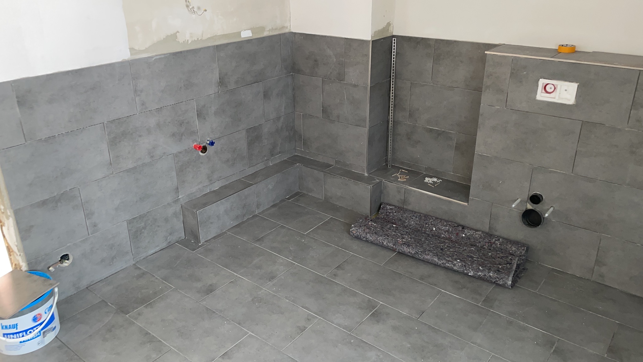 Szare, prostokątne płytki ceramiczne ułożone na ścianach i podłodze w trakcie prac wykończeniowych w łazience, widoczne otwory na instalacje i wiadro z zaprawą Knauf.