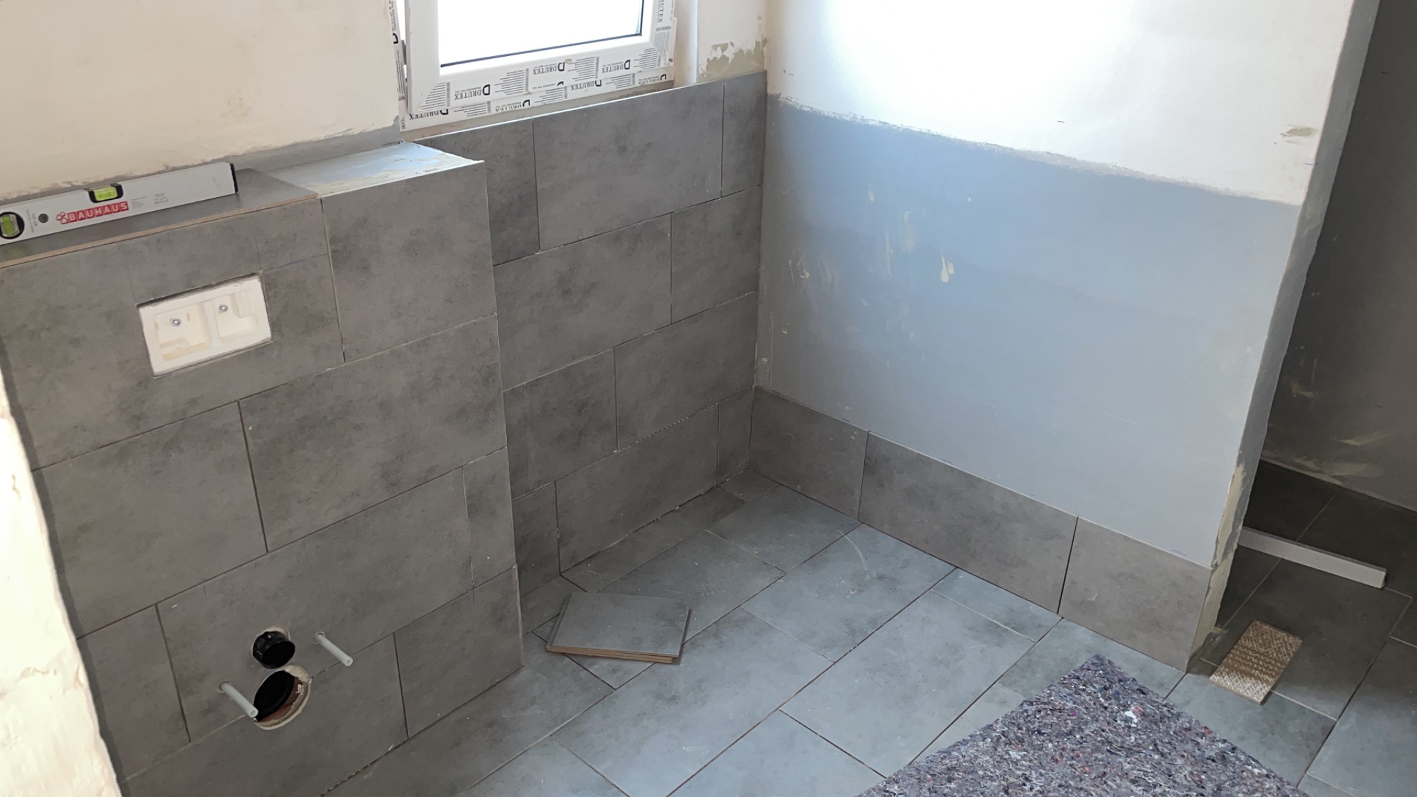 Szare płytki ceramiczne ułożone na ścianie i podłodze w trakcie prac wykończeniowych w łazience w Ostrowie Wielkopolskim, widoczny poziomica, fragment ściany przygotowany do malowania.