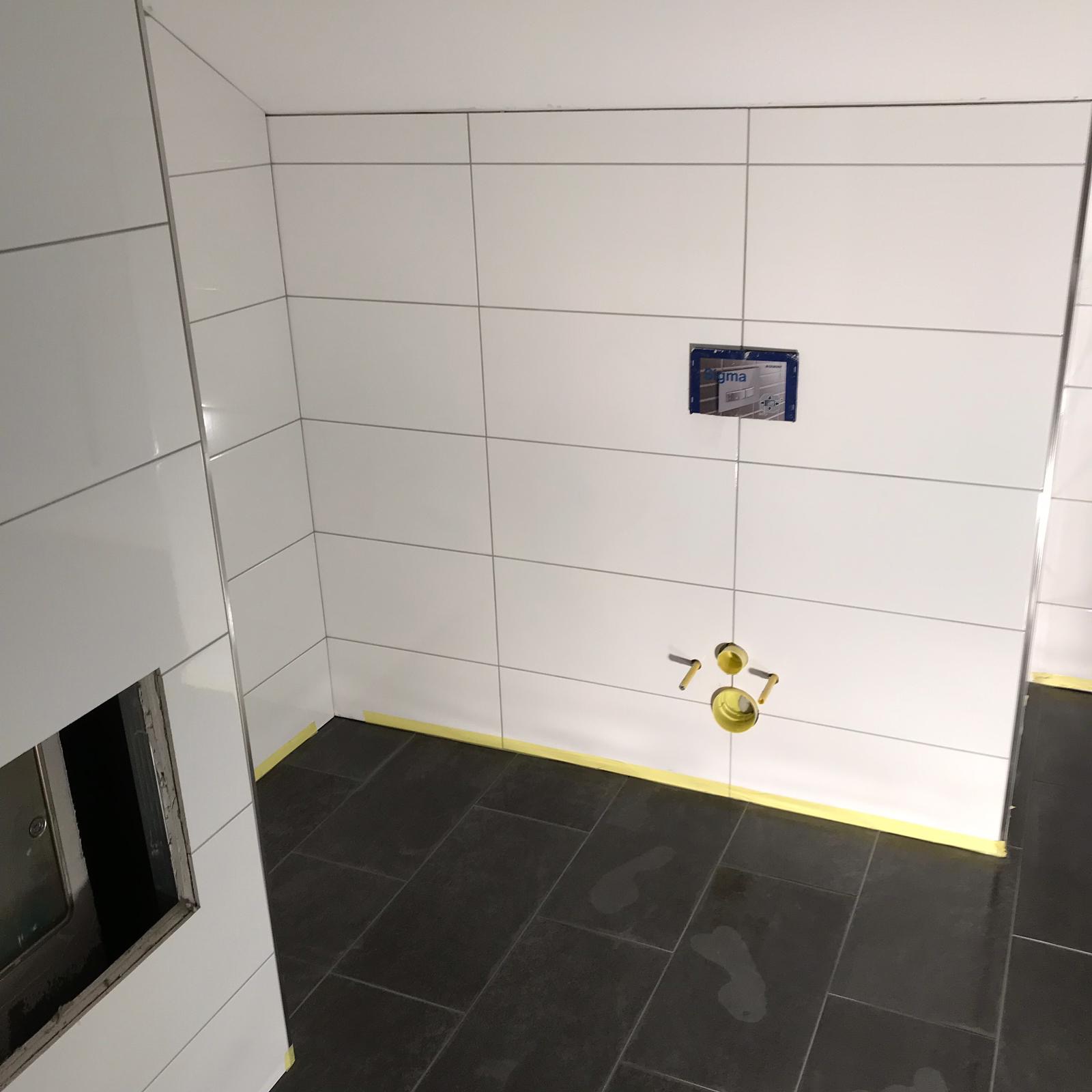 Świeżo wykafelkowana łazienka z białymi płytkami i ciemną podłogą, zabezpieczona żółtą taśmą malarską, widoczne otwory na armaturę i zabudowa podtynkowego systemu spłukiwania.