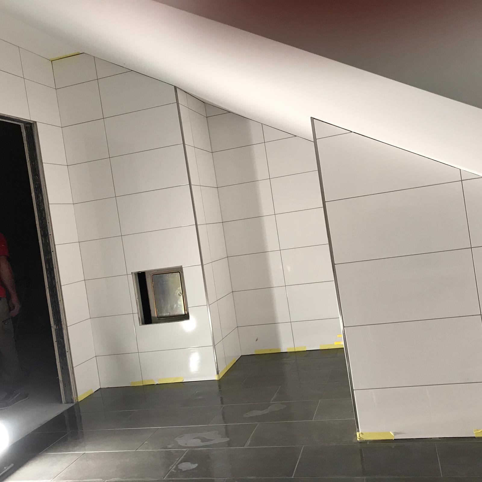 Niedokończona łazienka z białymi płytkami ściennymi i ciemnoszarą podłogą, zabezpieczona żółtą taśmą malarską przy podłodze i w narożnikach, widoczny właz rewizyjny.